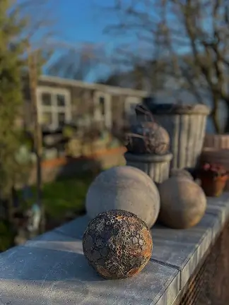 Række af dekorative sten og krukker på en hvid mur i en have med drivhus i baggrunden.
